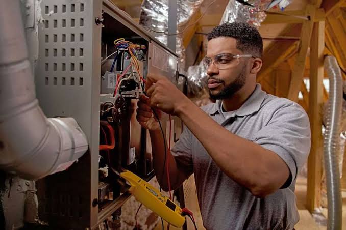 Technician checking the wiring of an indoor furnace beneath connected ducts